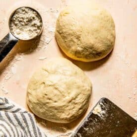 Two balls of dough on a floured surface with a metal measuring cup containing flour, a striped cloth, and a dough scraper nearby.