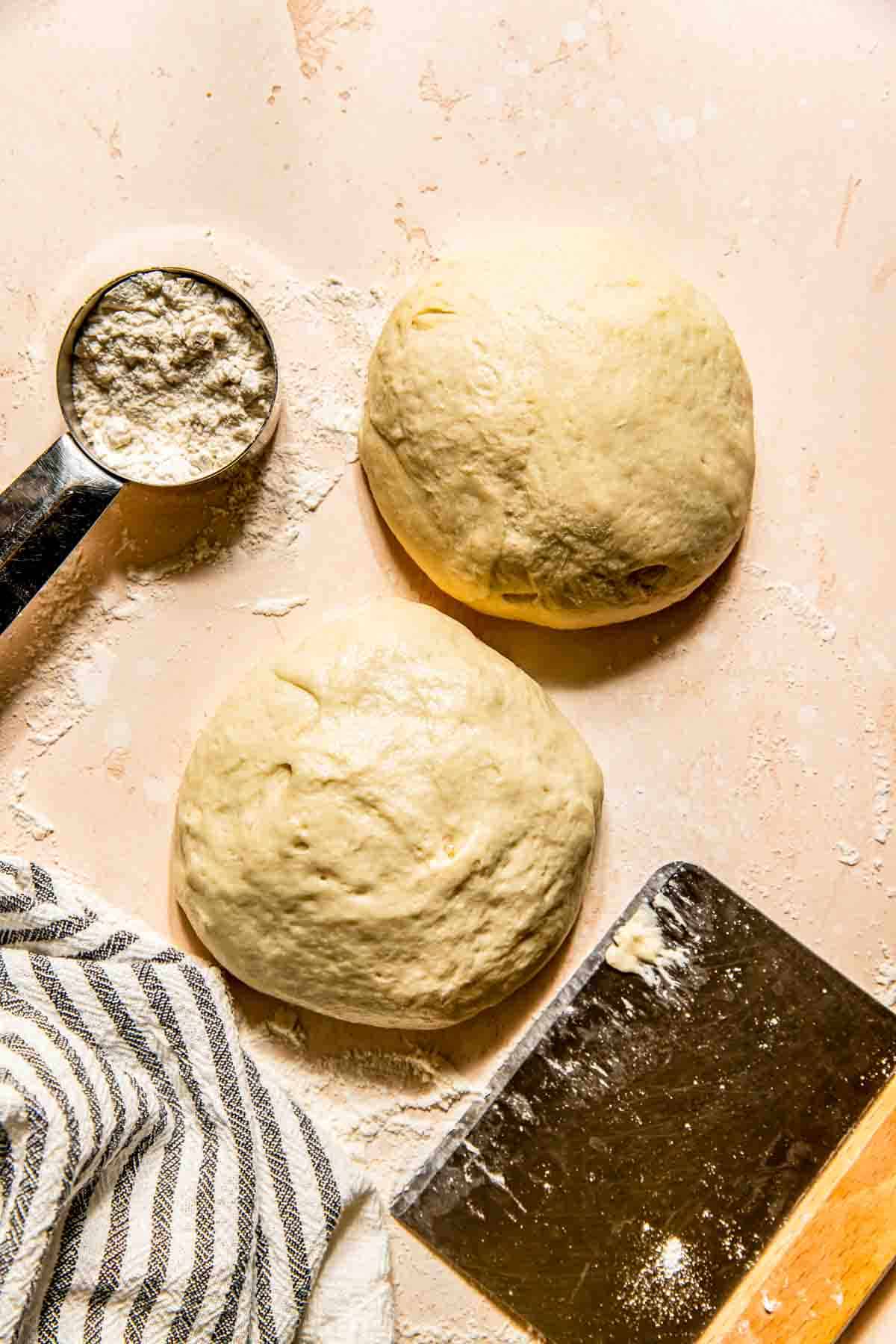 Two balls of dough on a floured surface with a metal measuring cup containing flour, a striped cloth, and a dough scraper nearby.
