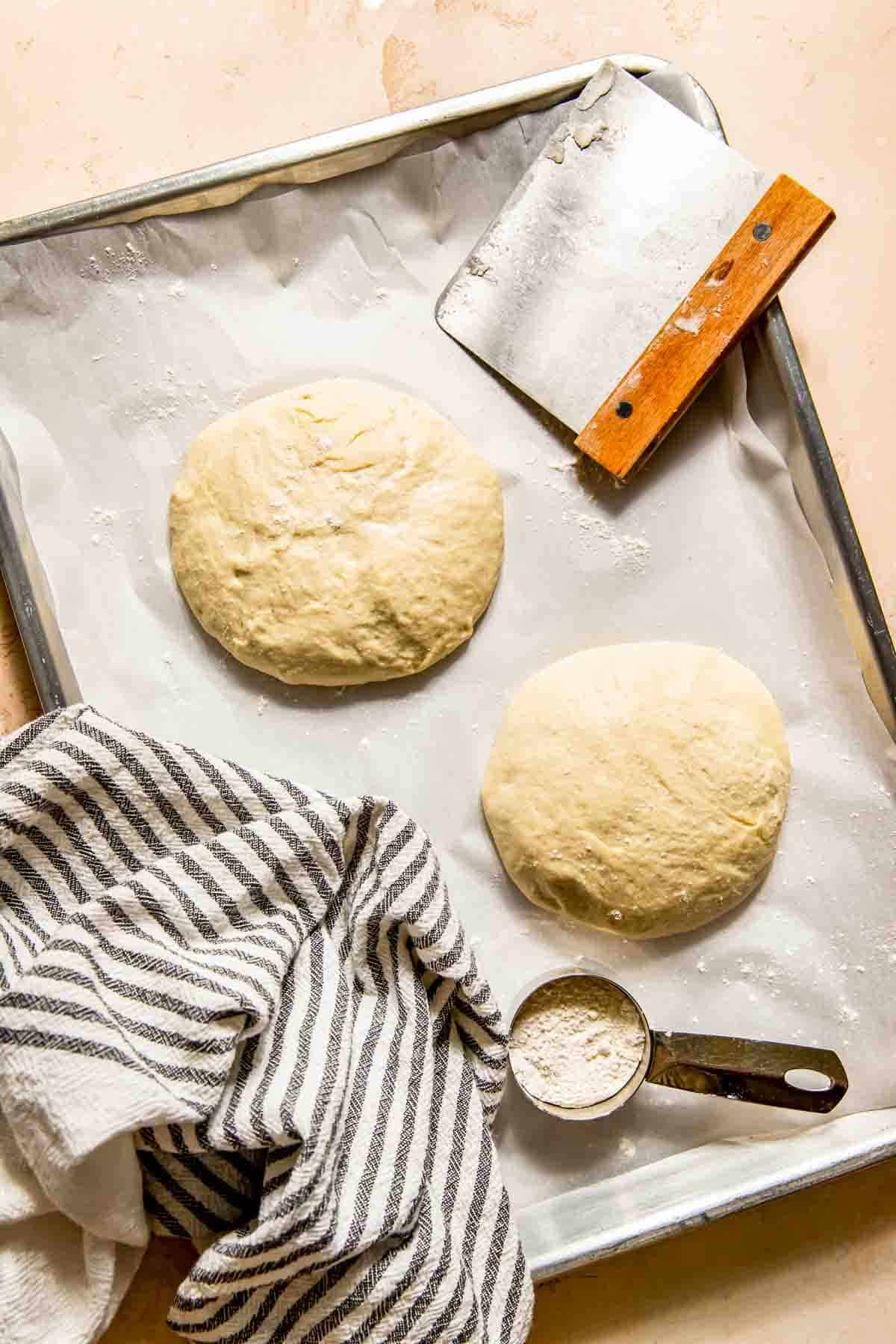 Two dough balls on a baking sheet with parchment paper, a dough scraper, a striped cloth, and a measuring cup with flour.