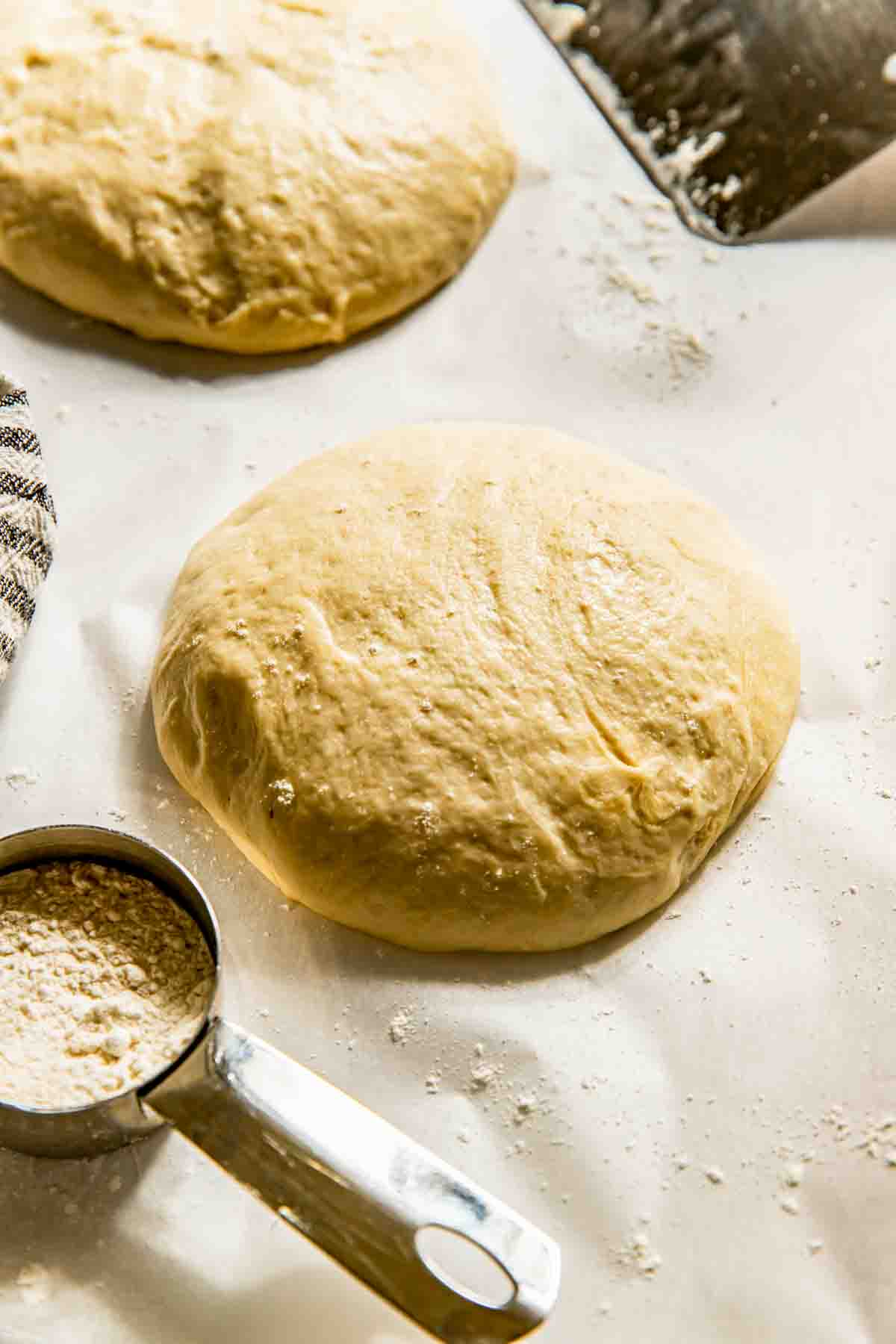 Balls of raw dough on a floured surface with a metal measuring cup filled with flour nearby.