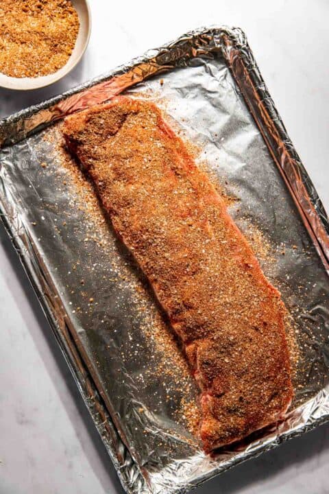 Raw rack of ribs seasoned with spices on a foil-lined baking sheet, next to a bowl of seasoning.