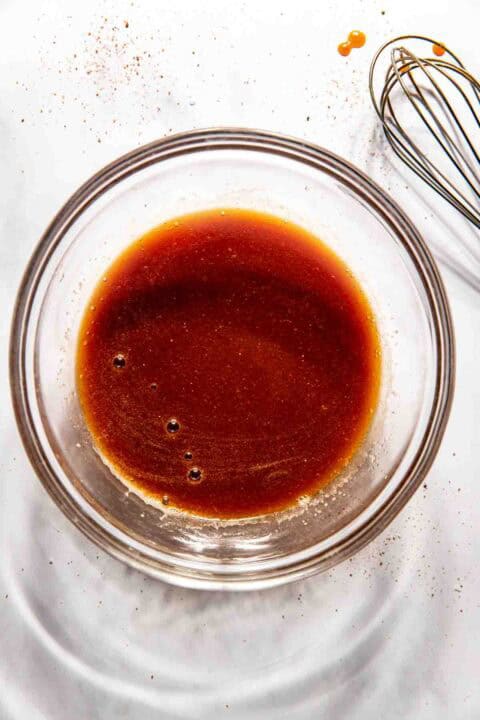A glass bowl containing a bbq sauce ingredients with a whisk beside it on a white surface.