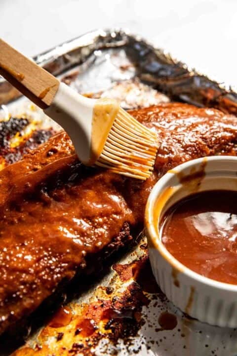 Brushing barbecue sauce on a rack of ribs on foil, with a small bowl of sauce nearby.