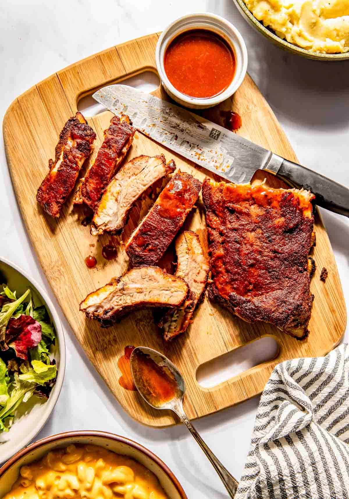 Sliced baby back ribs on a wooden board with a knife, small bowl of sauce, and spoon, surrounded by dishes of mashed potatoes and salad.