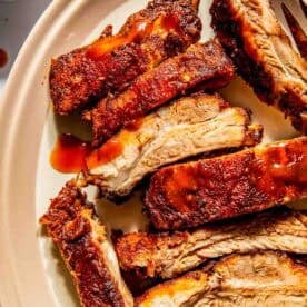 Close-up of sliced, seasoned baby back ribs on an oval plate with barbecue sauce. A small cup of sauce is visible in the background.