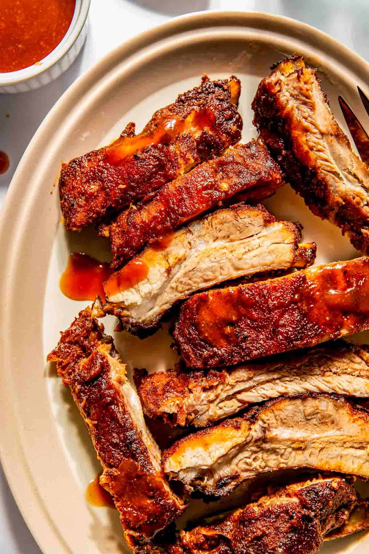 Close-up of sliced, seasoned baby back ribs on an oval plate with barbecue sauce. A small cup of sauce is visible in the background.