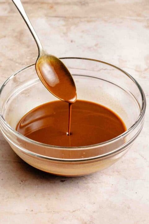 A spoon drizzling roux into a glass bowl on a marble surface.