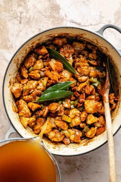 A pot of cooked chicken with vegetables and bay leaves, seasoned with spices. A glass measuring cup is pouring in broth.