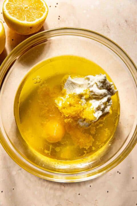Glass bowl with egg, lemon zest, ricotta, oil, and other ingredients. Halved lemons are beside the bowl on a marble surface.