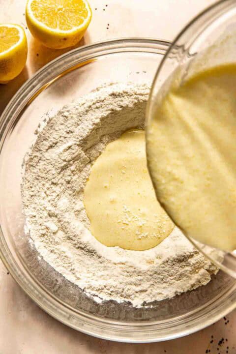 Batter is being poured into a well in a bowl of flour, with two halved lemons nearby.