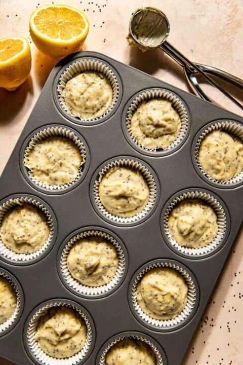 A muffin tray with twelve paper liners filled with uncooked lemon chia seed batter. A lemon and a scoop with batter are nearby on the countertop.