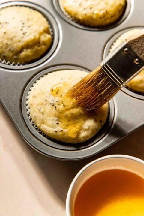 A brush applies honey to freshly baked muffins in a muffin tin, with a cup of honey nearby.