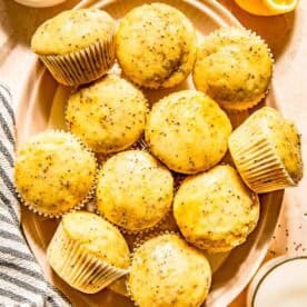 A plate of lemon chia seed muffins is surrounded by a halved lemon, a small dish of honey with a honey dipper, and a glass of milk.