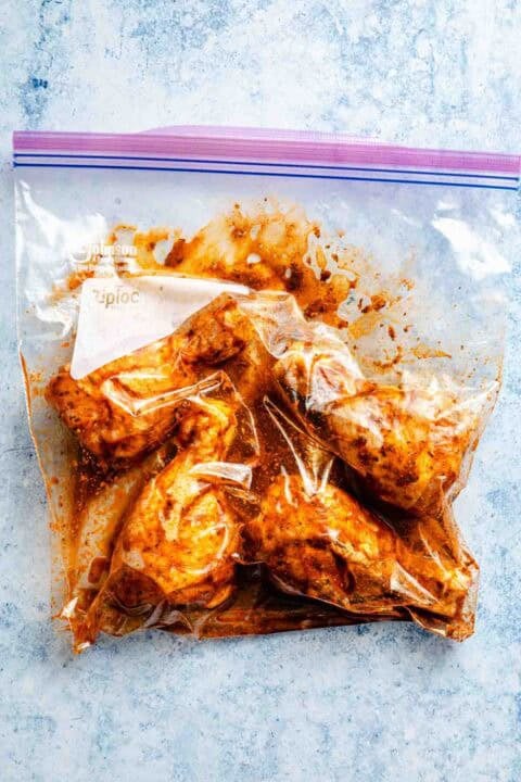Raw chicken drumsticks marinating in a sealed plastic bag on a textured blue surface.