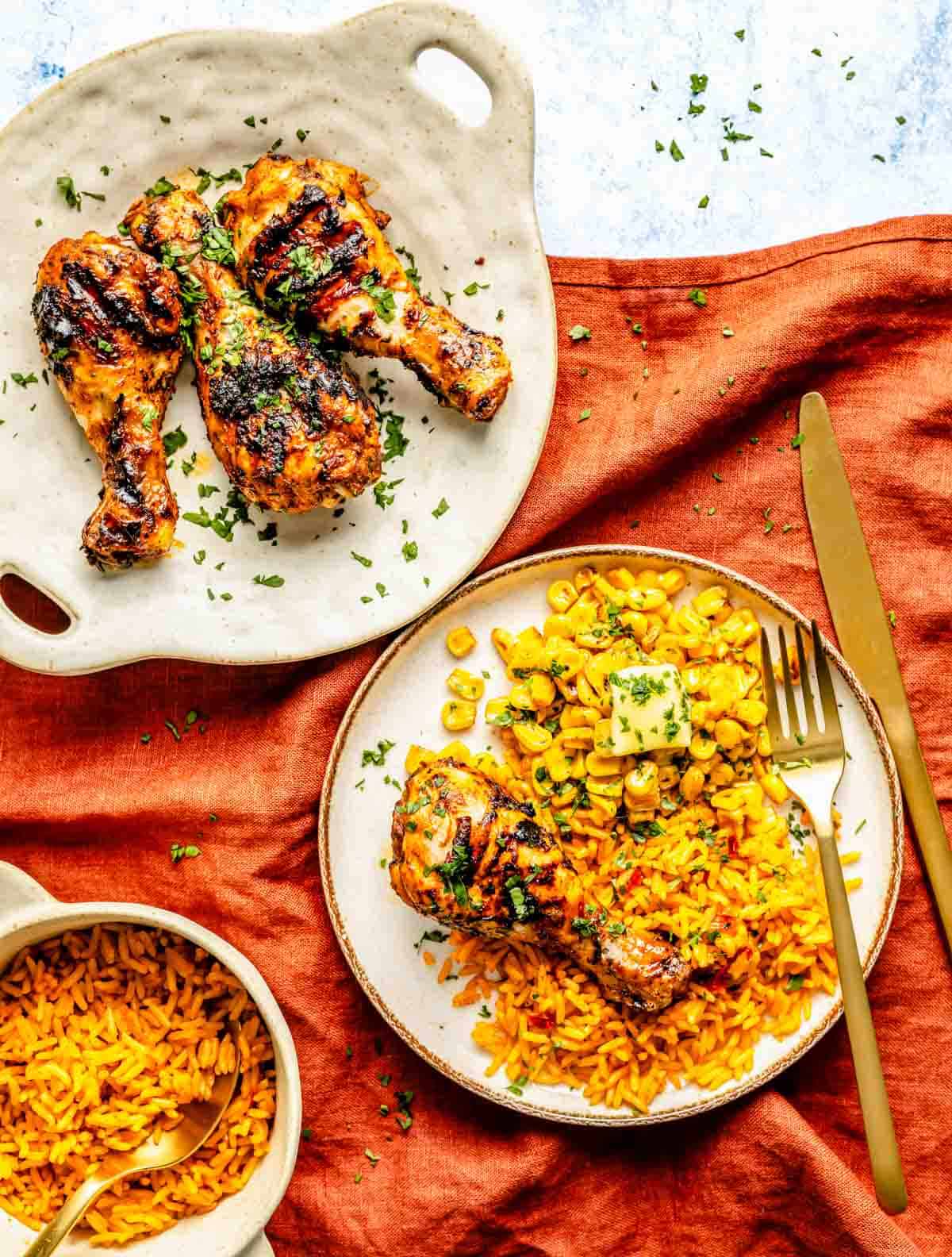 Grilled chicken drumsticks on a plate with corn and rice, garnished with herbs, alongside a bowl of rice.