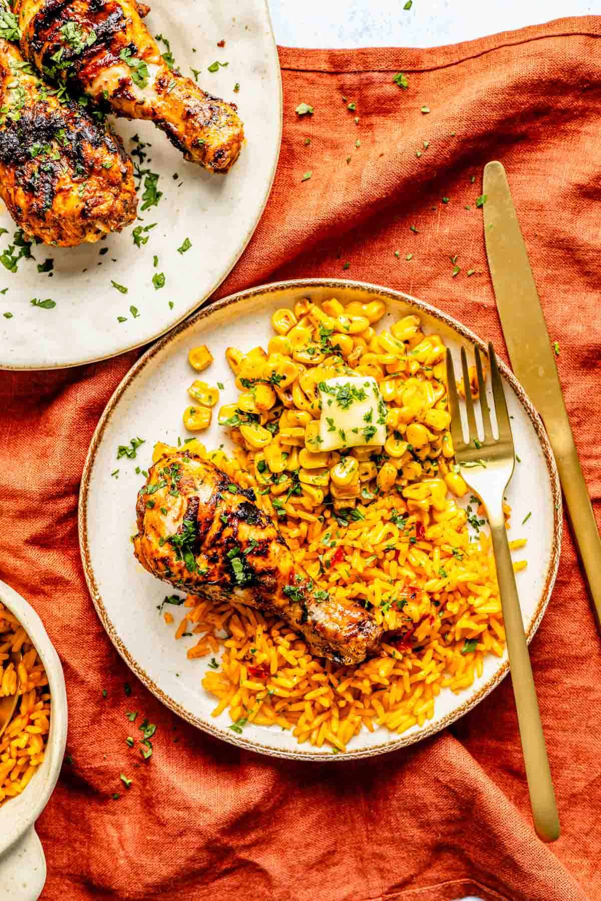 Grilled chicken drumsticks and yellow rice with corn on a white plate, garnished with parsley, placed on an orange cloth. Gold fork and knife are beside the plate.