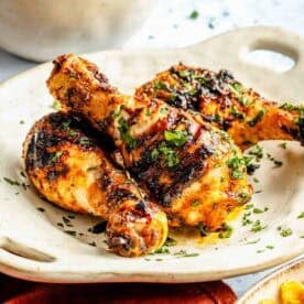Grilled chicken drumsticks garnished with herbs on a white plate, with a side of corn and a bowl of rice in the background.