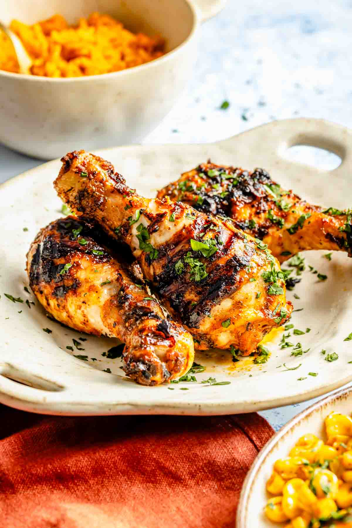 Grilled chicken drumsticks garnished with herbs on a white plate, with a side of corn and a bowl of rice in the background.