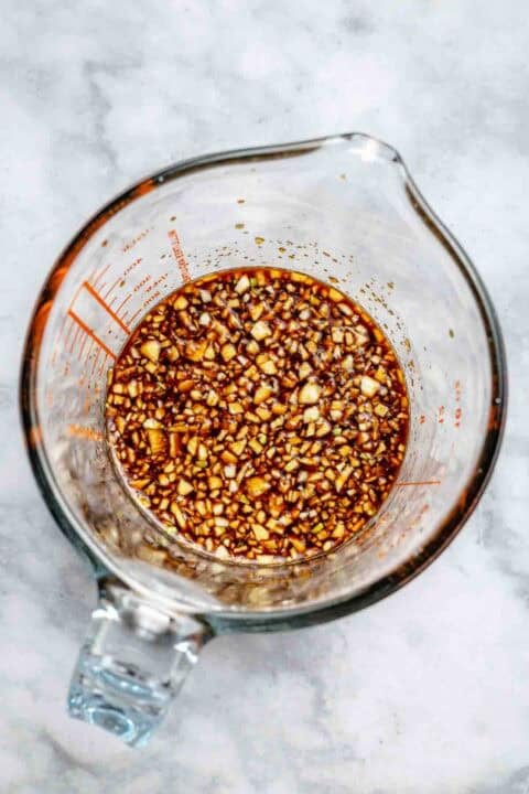 A glass measuring cup filled with a marinade containing chopped garlic and soy sauce, placed on a marble surface.