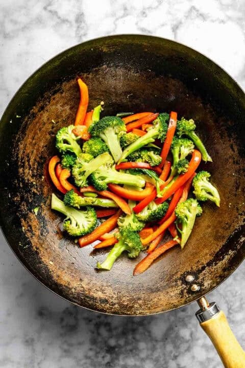 Stir-fried broccoli and red bell pepper slices in a large wok on a marble surface.