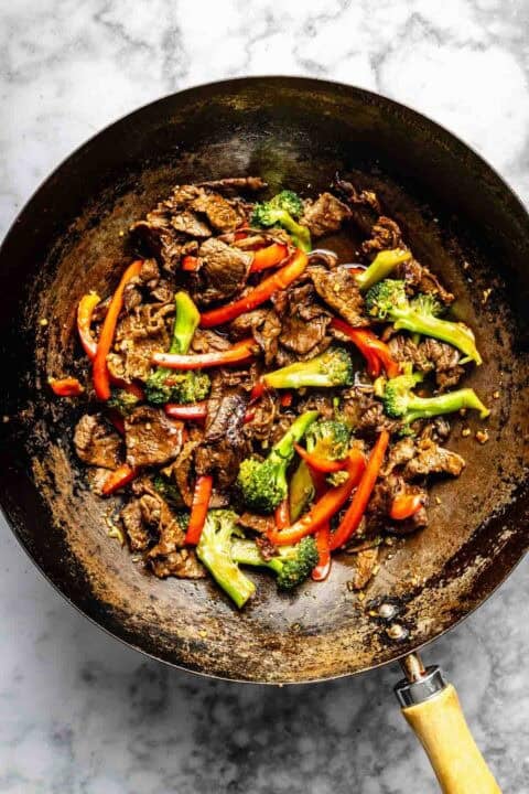 A wok filled with stir-fried beef slices, red bell peppers, and broccoli on a marble surface.