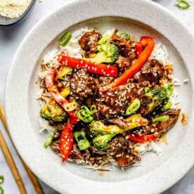 A bowl of rice topped with stir-fried beef, broccoli, red bell peppers, and sesame seeds, garnished with sliced green onions. Chopsticks lie beside the bowl.
