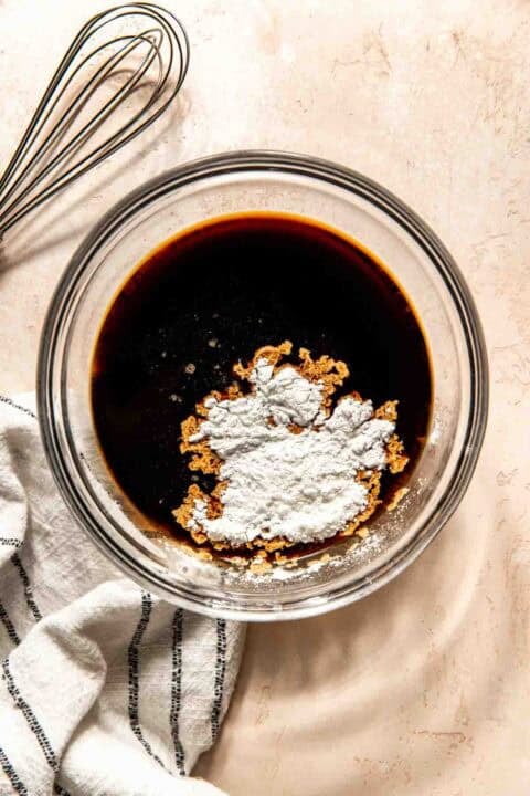 A glass bowl with soy sauce mixture and cornstarch is on a counter next to a metal whisk and striped cloth.