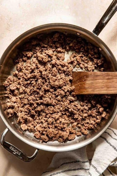 Cooked ground beef in a stainless steel pan with a wooden spatula, placed on a light-colored surface with a striped cloth nearby.