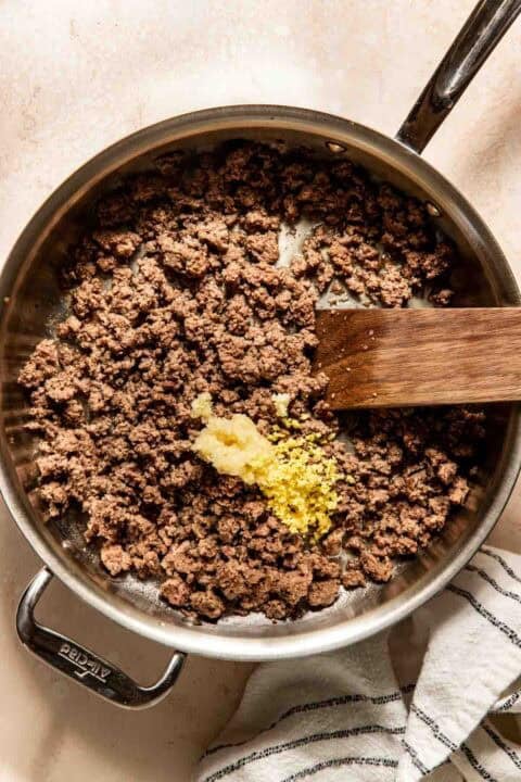 Ground beef cooking in a skillet with minced garlic and ginger, shown with a wooden spatula and a kitchen towel nearby.