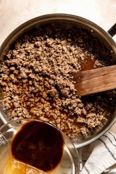 Ground beef browning in a skillet with a wooden spatula, next to a measuring cup containing sauce ingredients being poured in.