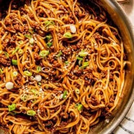 A pan of stir-fried noodles with minced meat, topped with sliced green onions and sesame seeds.