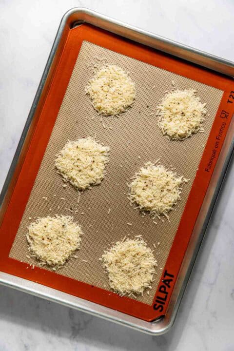 Six mounds of grated parmesan cheese on a silpat-lined baking sheet.