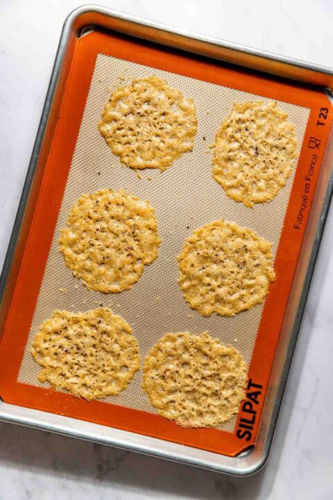 Six baked parmesan cheese crisps on a baking sheet lined with a silicone mat.