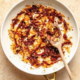 A white frying pan with sautéed onions and sun-dried tomatoes, and a wooden spoon on a beige surface.