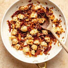 A skillet with cooked chicken, sun-dried tomatoes, and onions, stirred with a wooden spoon on a beige surface.