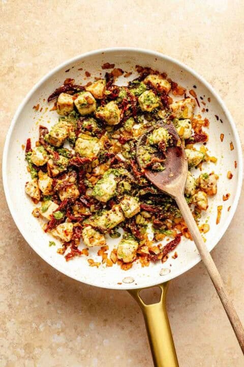 A pan filled with cooked chicken, mixed with sun-dried tomatoes, spices, and pesto, is being stirred with a wooden spoon on a beige background.