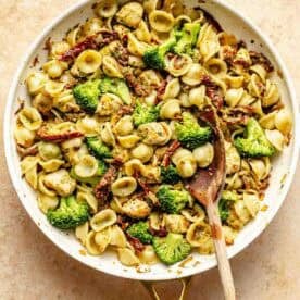 A skillet of orecchiette pasta with pieces of chicken, broccoli, and sun-dried tomatoes, stirred with a wooden spoon.