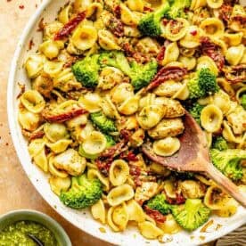 A skillet filled with orecchiette pasta, broccoli, sun-dried tomatoes, and chicken pieces. A wooden spoon is in the skillet. A small bowl of basil pesto is next to the skillet.