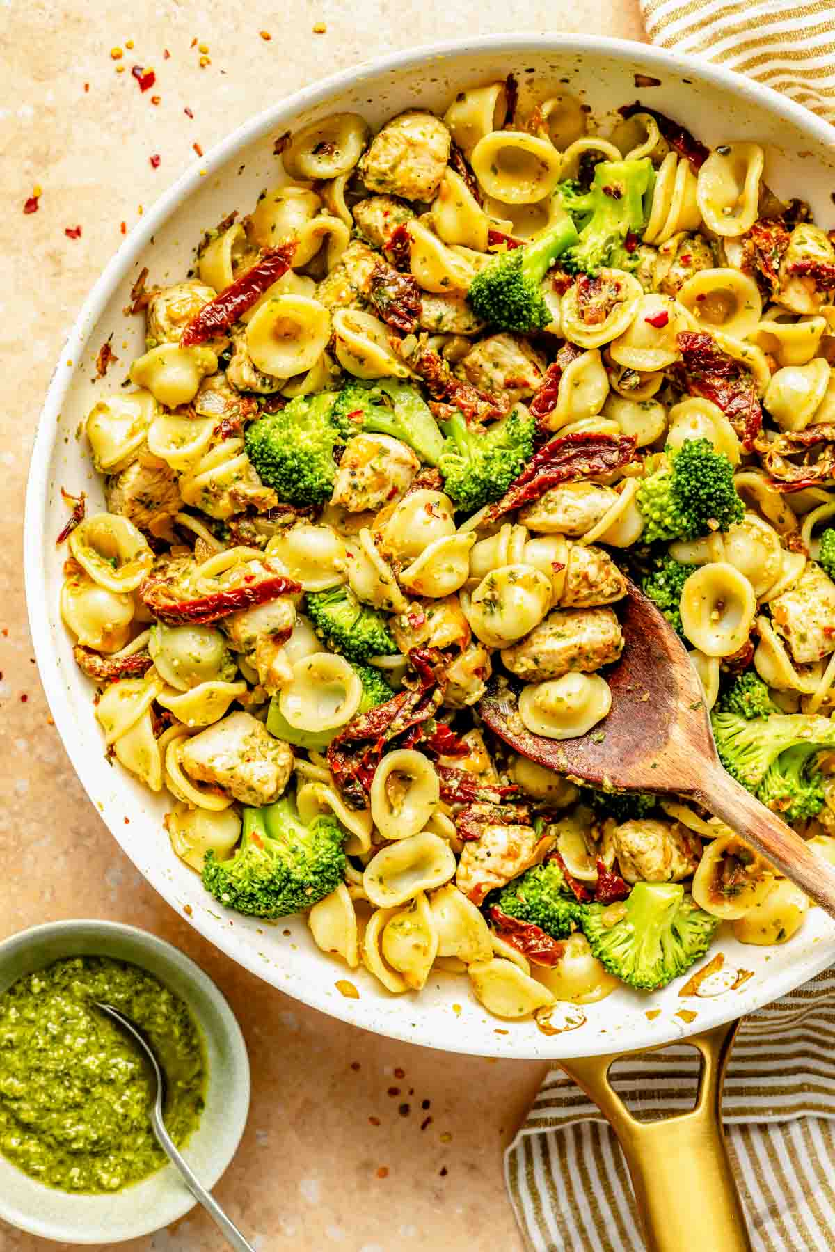 A skillet filled with orecchiette pasta, broccoli, sun-dried tomatoes, and chicken pieces. A wooden spoon is in the skillet. A small bowl of basil pesto is next to the skillet.