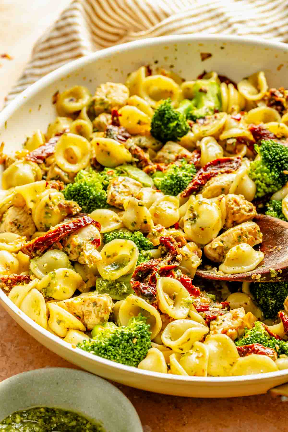 A bowl of orecchiette pasta with pieces of chicken, broccoli florets, and sun-dried tomatoes, tossed in a pesto sauce. A wooden spoon is partially visible in the dish.