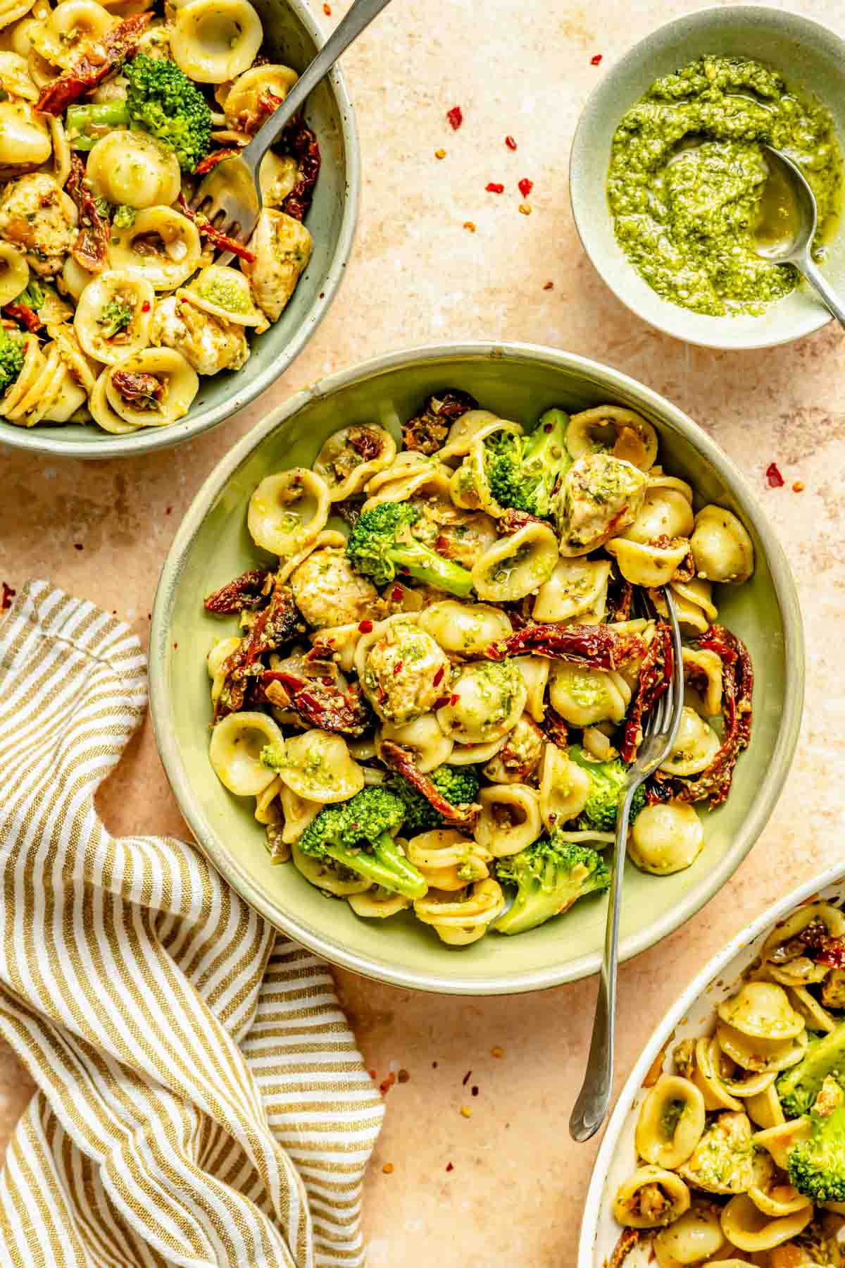 Bowls of orecchiette pasta with broccoli, sun-dried tomatoes, chicken, and pesto. A striped napkin and a bowl of pesto sit beside them.