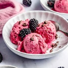 A bowl of bright blackberry ice cream with three scoops, topped with fresh blackberries and chocolate shavings, with a spoon on the side. Additional blackberries and another bowl are in the background.