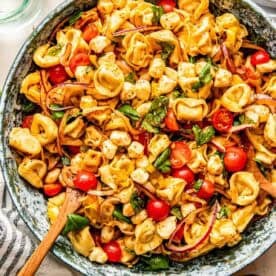 A bowl of tortellini pasta salad with cherry tomatoes, mozzarella balls, red onion, and fresh basil, mixed with a wooden spoon.
