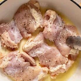 Raw, seasoned chicken thighs in a white pot with melted butter, shown with metal tongs, ready for cooking.