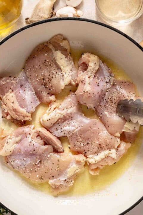 Raw, seasoned chicken thighs in a white pot with melted butter, shown with metal tongs, ready for cooking.