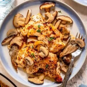 Creamy mushroom chicken served with a creamy mushroom sauce and garnished with fresh herbs on a round plate, accompanied by a fork and knife.