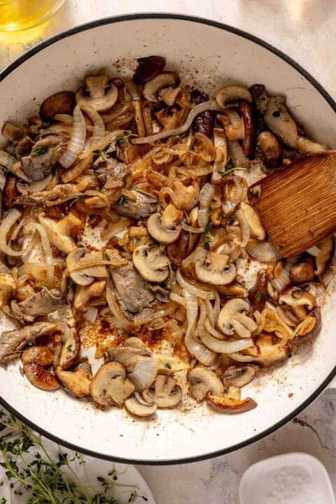 Sliced mushrooms and onions sautéing in a white pan with a wooden spatula, garnished with fresh herbs.