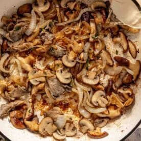 Sliced mushrooms and onions cooking in a white skillet, with white wine being poured in from a glass measuring cup.