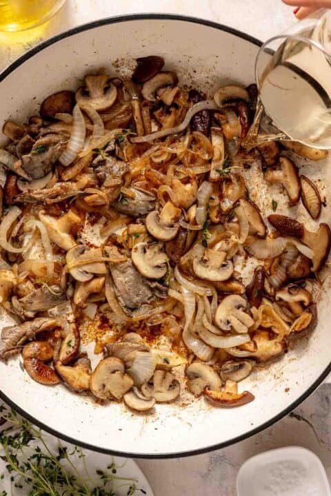 Sliced mushrooms and onions cooking in a white skillet, with white wine being poured in from a glass measuring cup.