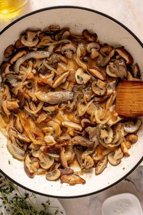 Sautéed mushrooms and onions in a white skillet with a wooden spatula.